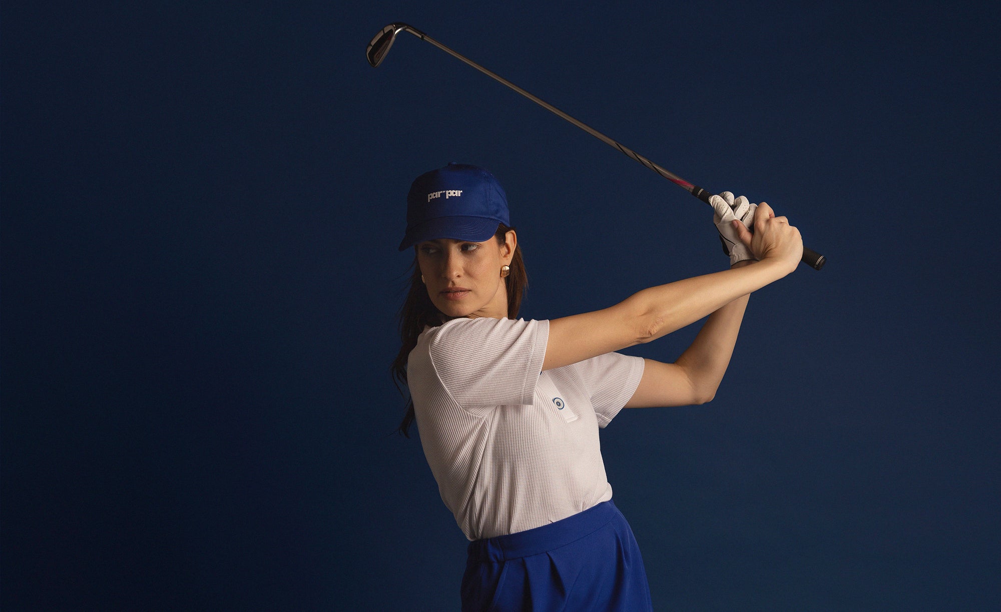 Woman swinging a golf club wearing Par by Par blue cap and white polo